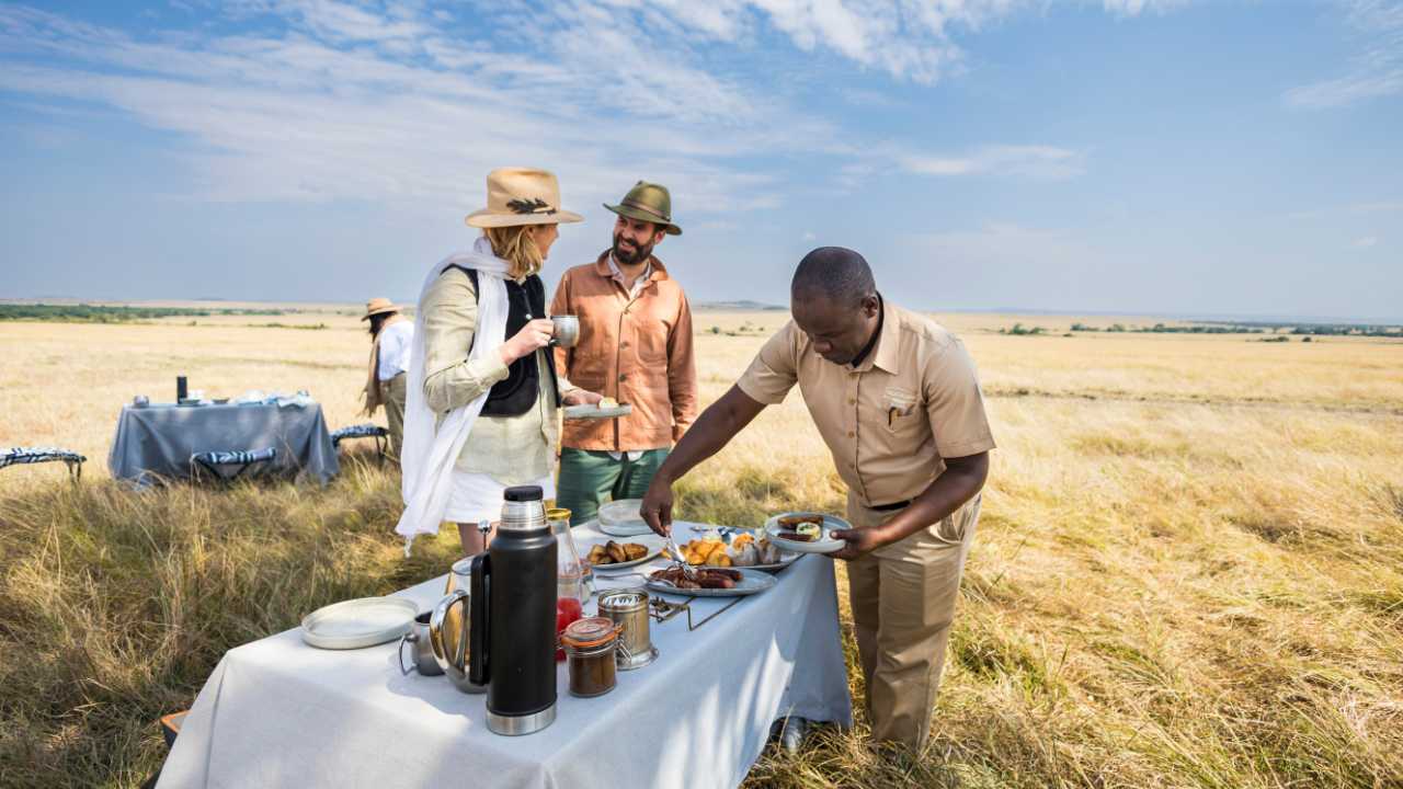 Three guests are enjoying a luxury snack in the Maasai Mara plains, Kenya, with a host.