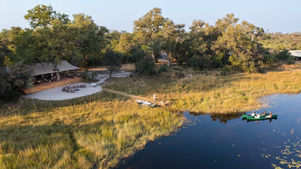 Aerial view of the main lodge of Explorers Camp in the Okavango Delta, Botswana. Calm waterways in the front with two canoes, in the backdrop is the main guest lodge with lush grass in front of the guest lodge.