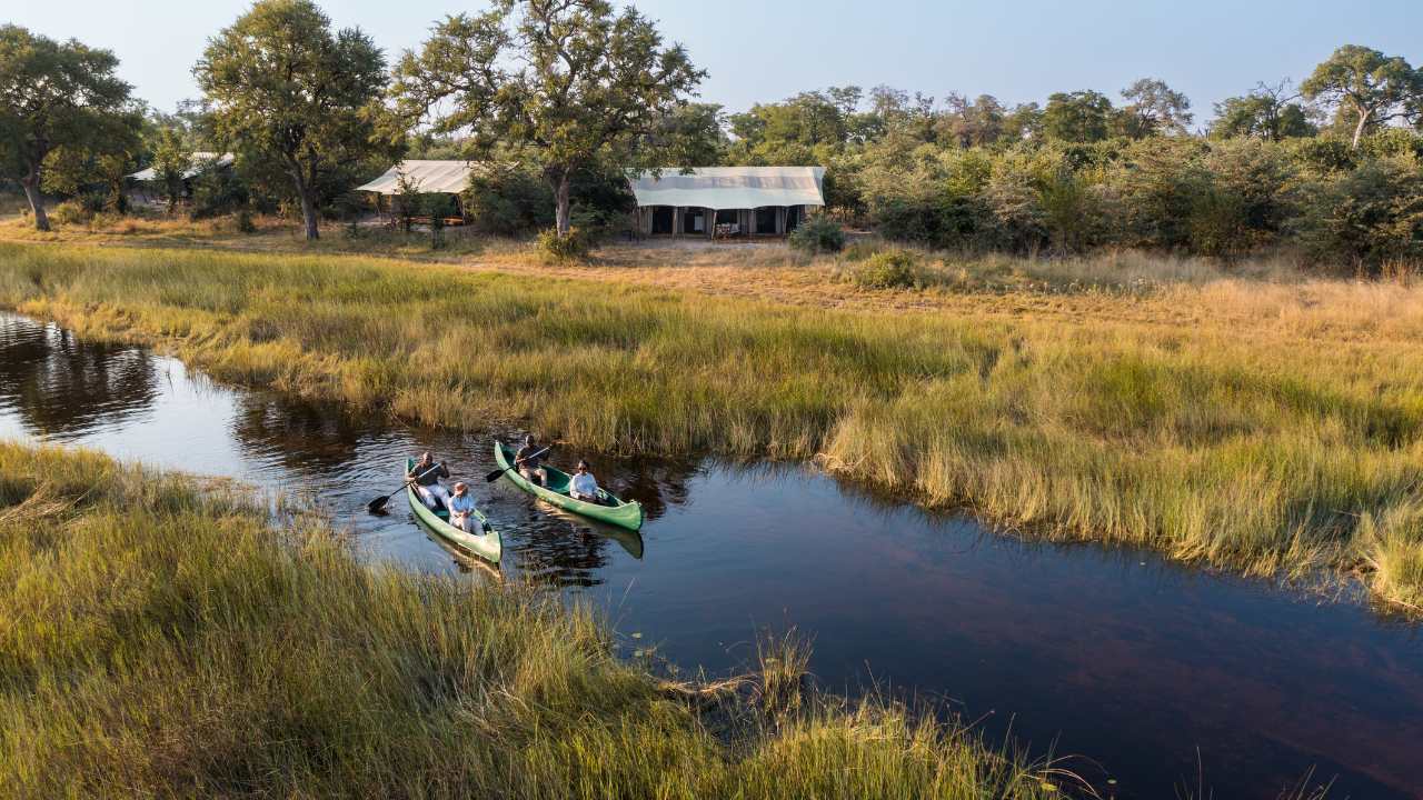 Aerial shot of a quiet river in the Okavango in the foreground, with two guides taking guests out on a canoe safari on the Okavango Delta in Botswana. Canoes and river in the foreground, golden grass in the background, and right at the back, the Okavango Explorers Camp.