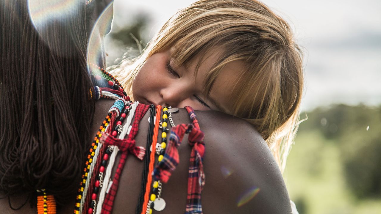 Kenyan safari guide in the bush with a child asleep on her shoulder, viewed from behind.