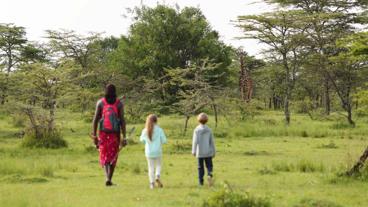 Kenyan safari guide in traditional attire leading children on a walking safari in the bush.