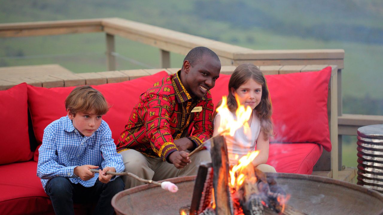 Kenyan safari host toasting marshmallows with a boy and girl at a campfire on a deck.