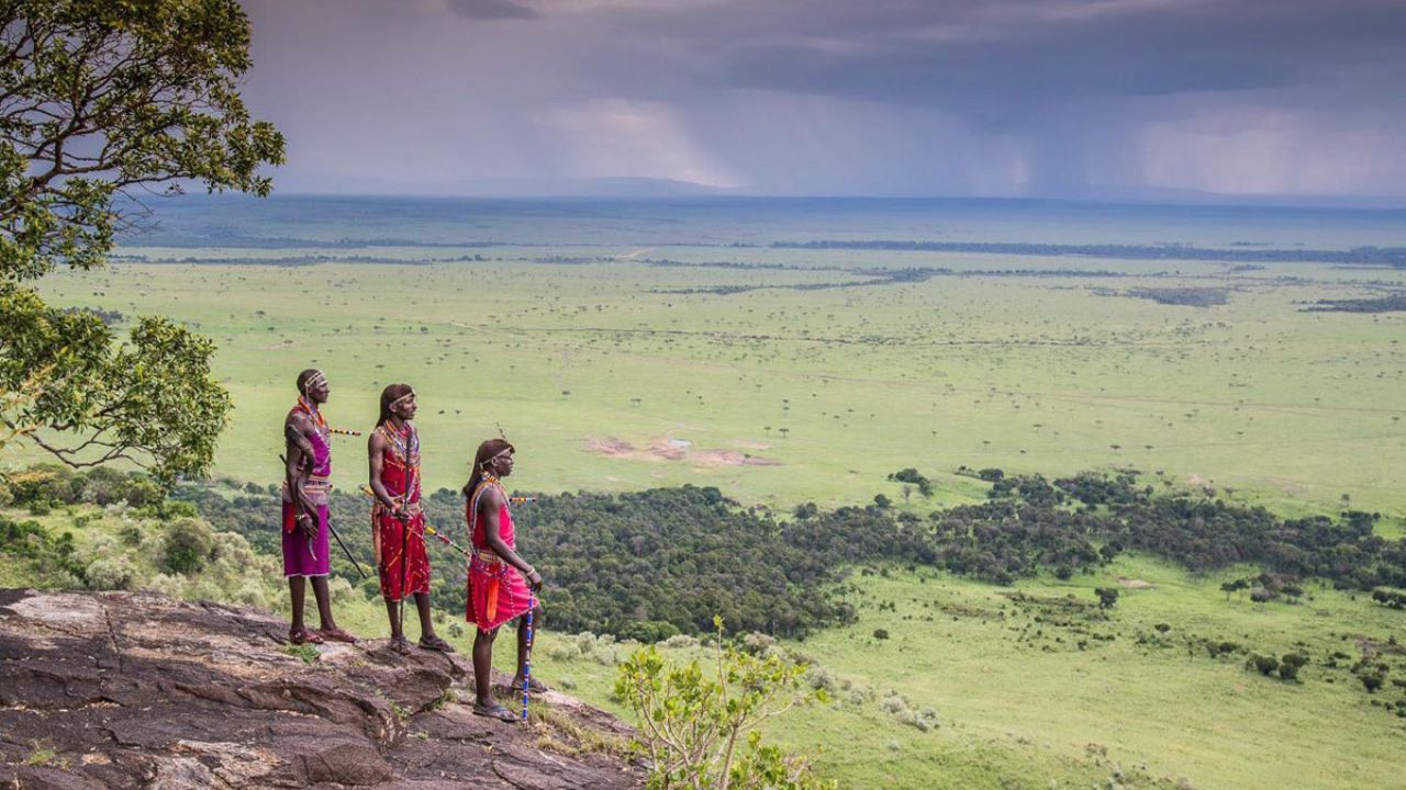 Three Maasai tribesmen standing on rock overlooking Maasai Mara plains, Kenya, African safari and cultural experience.