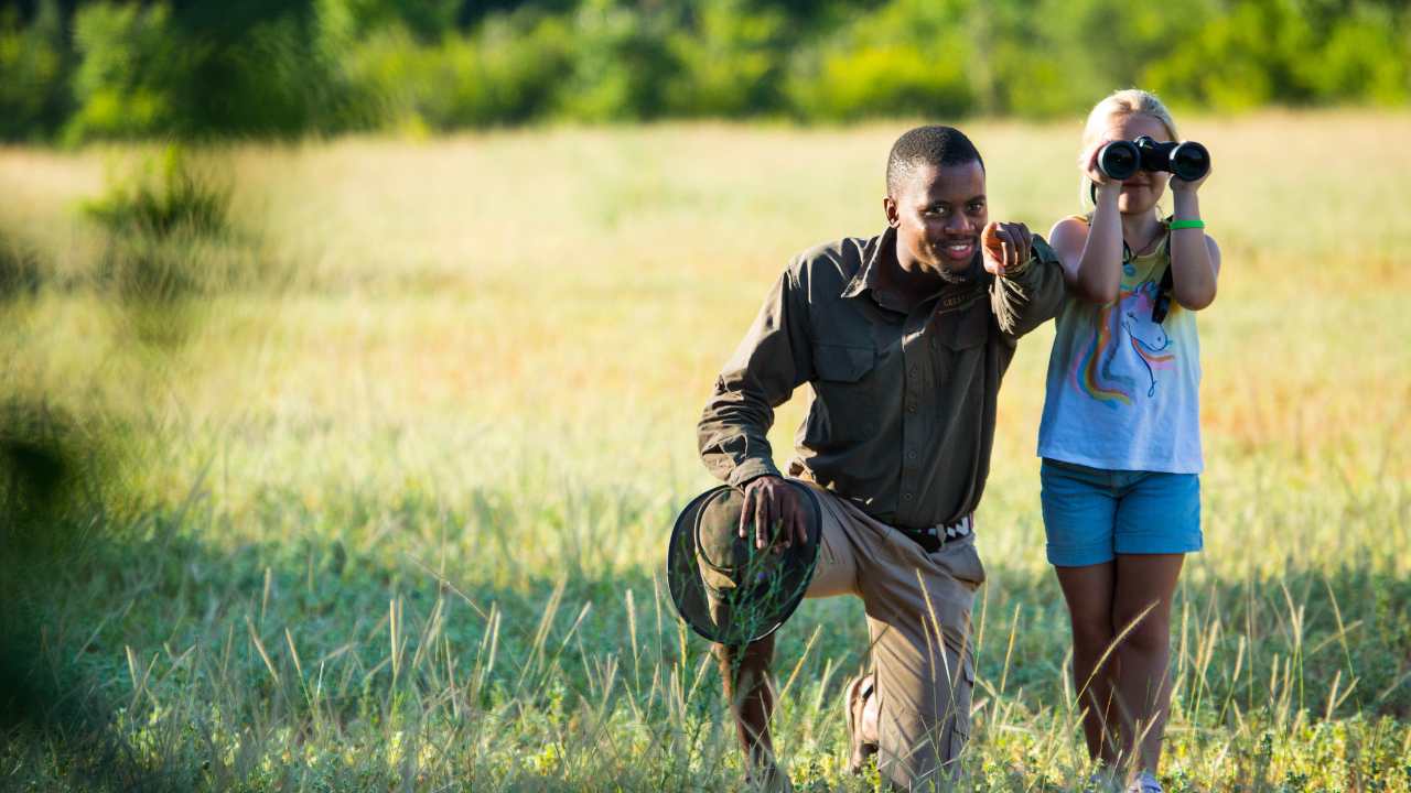 Game ranger points out wildlife to a young girl using binoculars on a family safari in the Okavango Delta, Botswana.