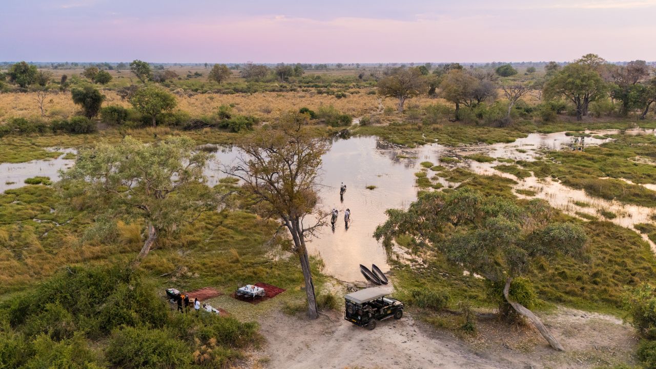 Aerial view of three traditional canoes arriving for sundowner drinks at Duba Plains Camp, with safari vehicle in foreground, Okavango Delta, Botswana.