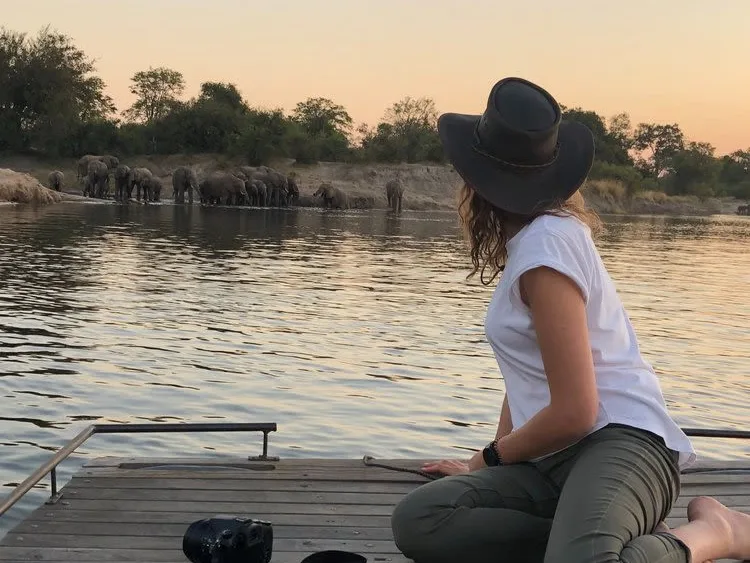 Woman on small wooden raft looking over Zambezi River at herd of elephants on the other side of the river, woman wearing white top and safari hat on a wildlife safari in Zambia.