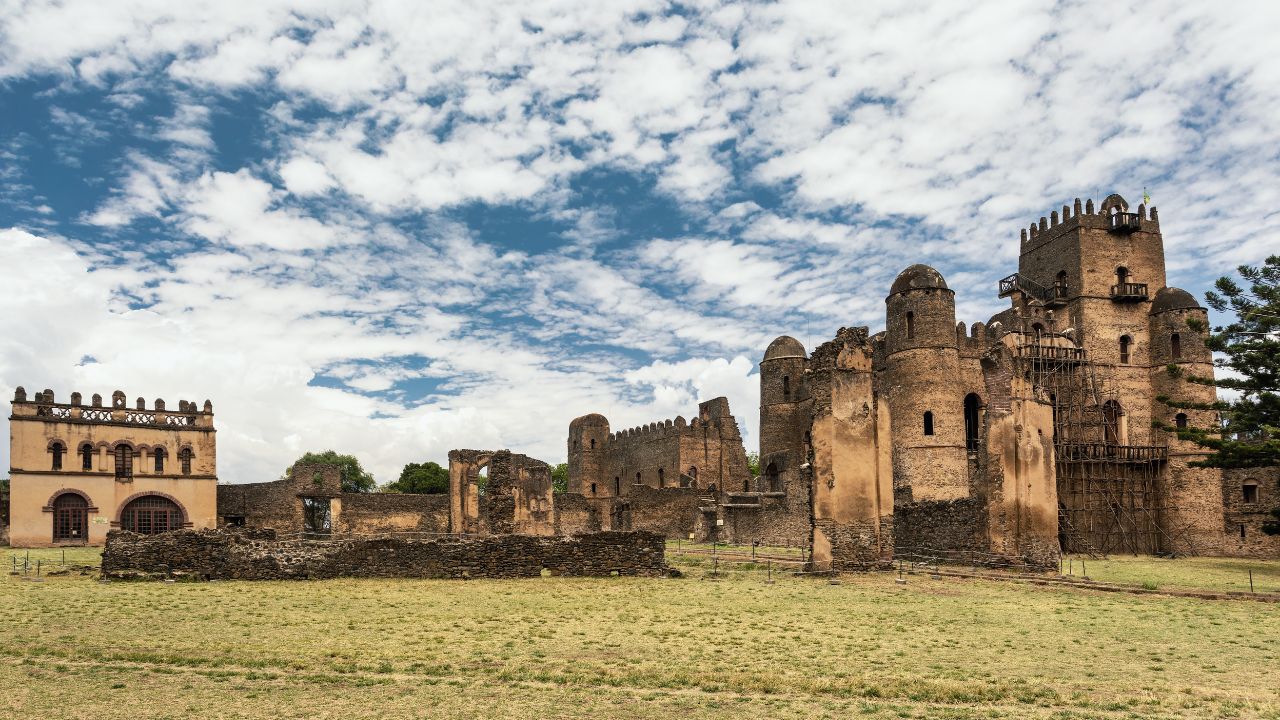 Wide-angle shot of Gondar castle with clouds, blue sky, and large grassy patch in front.