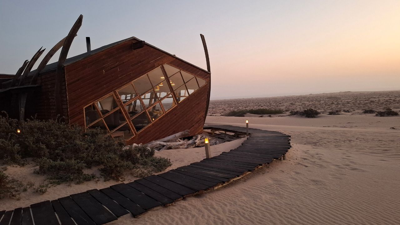 Main lodge of Shipwreck Lodge with black wooden walkway, lodge on left, desert on right