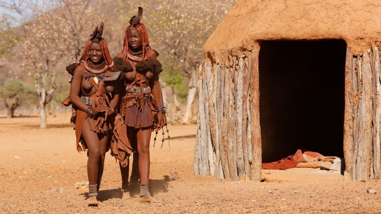 Two Himba ladies in full traditional dress with red braided clay hair, mud hut on right