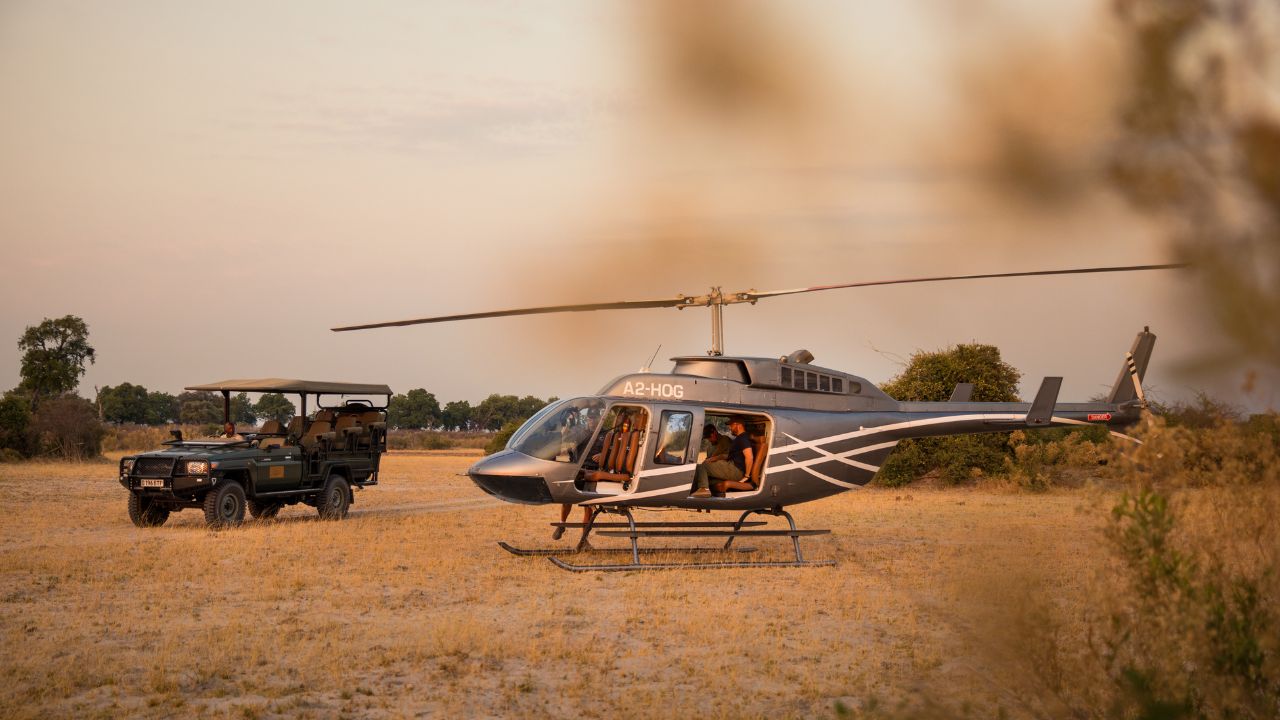 Guests returning from a helicopter safari at Sitatunga Private Island with sunset over golden grass and safari vehicle ready, Botswana.