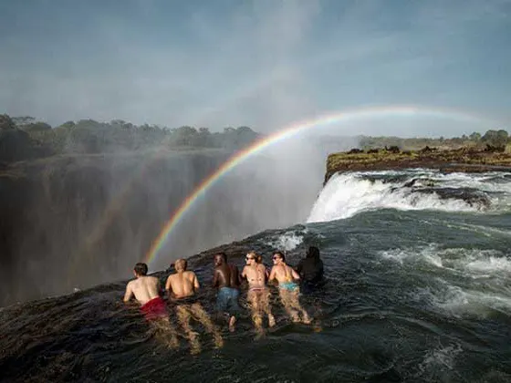 Devil's Pool in Zambia, group of six people in Devil's Pool in the Zambezi River overlooking Victoria Falls with rainbow from the spray in background.