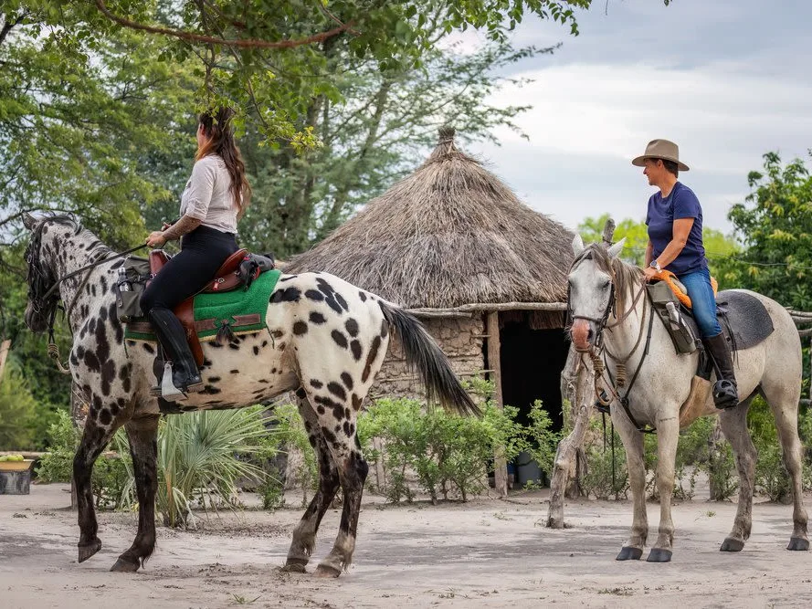 Two people on horseback, white horses with black spots, one man, one woman, in the background is an African mud hut with a straw roof, on a Zambian horseback safari.