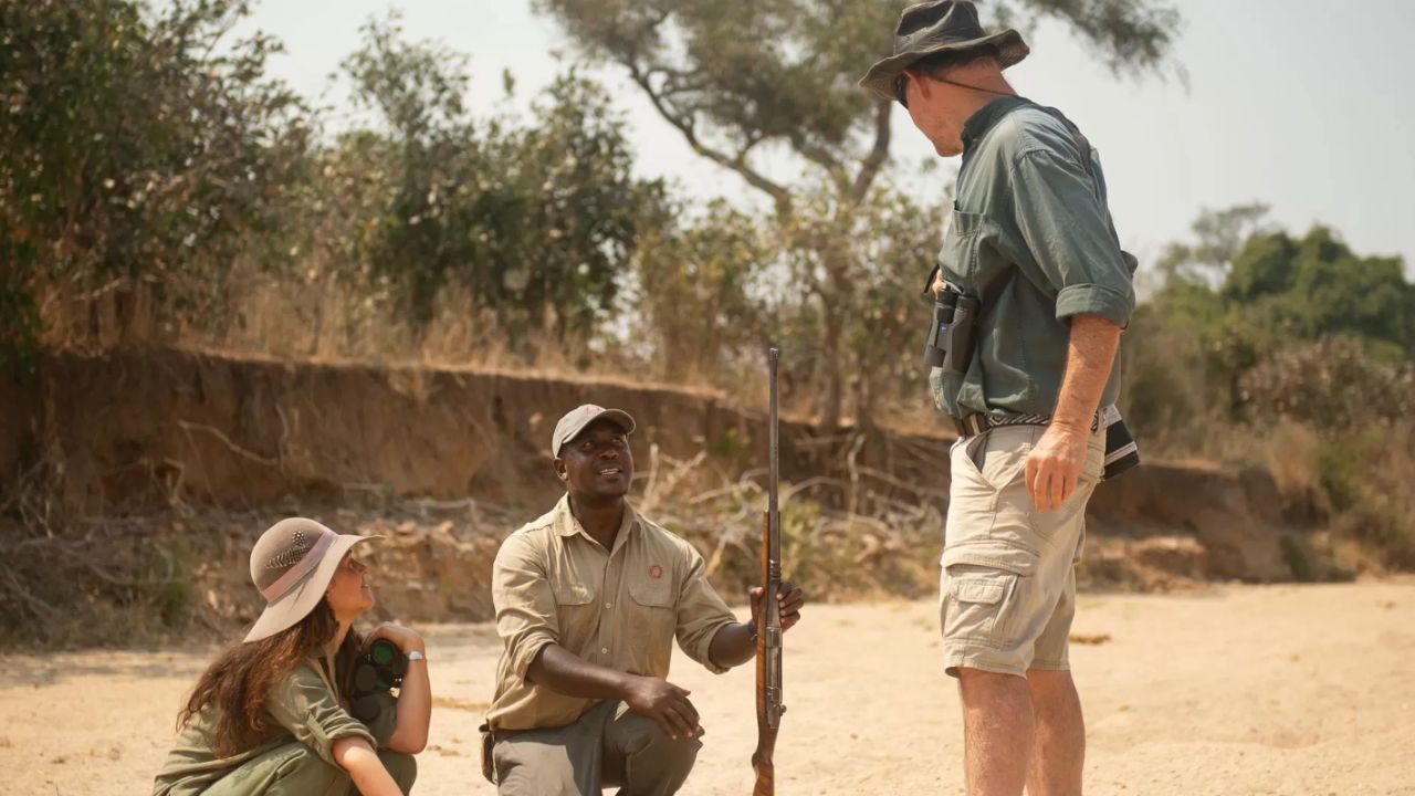 Safari ranger crouching in a sandy riverbed explaining animal tracks to two guests in the Serengeti.