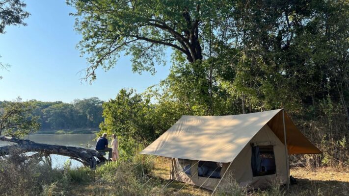 Canvas tent by the river with two guests seated on a log overlooking the Tanzanian bush.