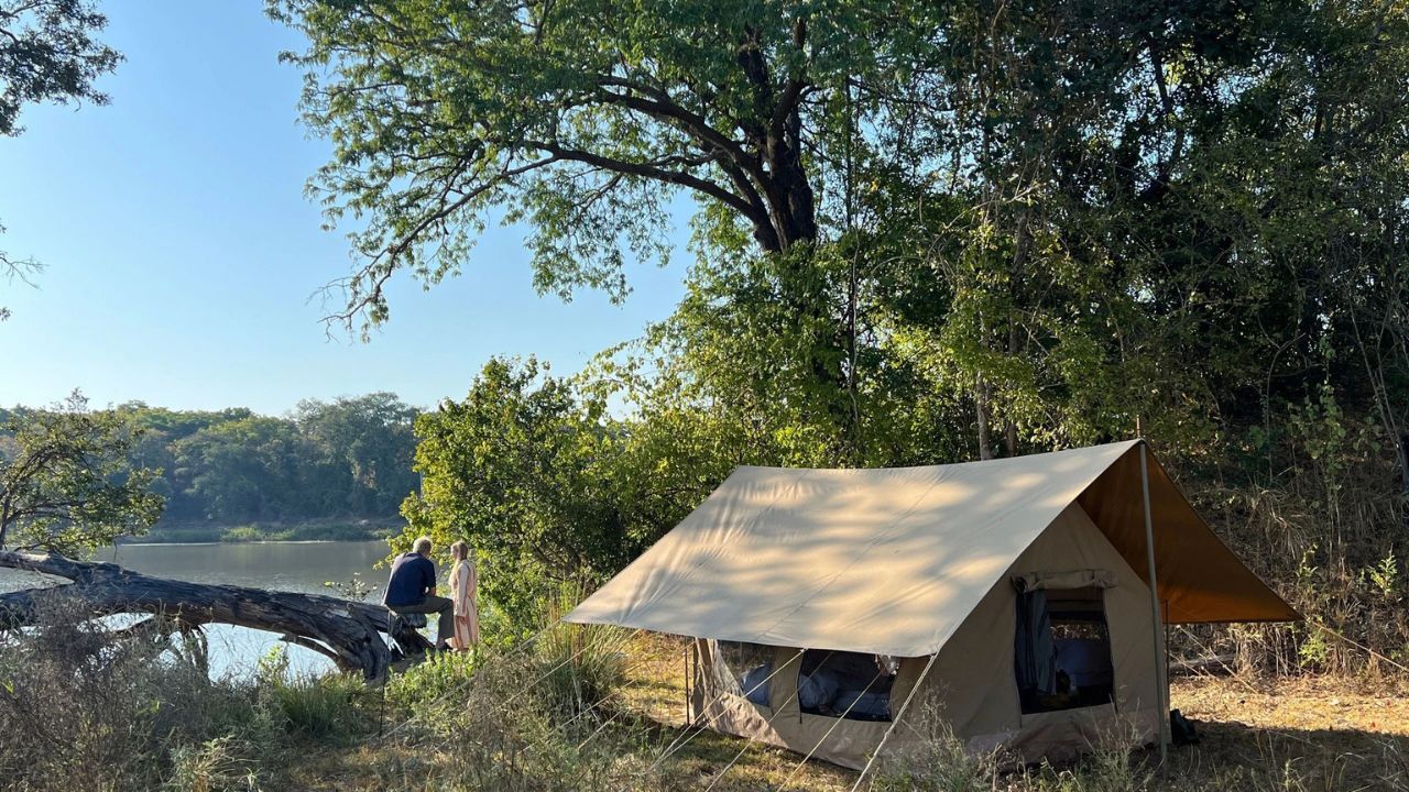 Canvas tent by the river with two guests seated on a log overlooking the Tanzanian bush.