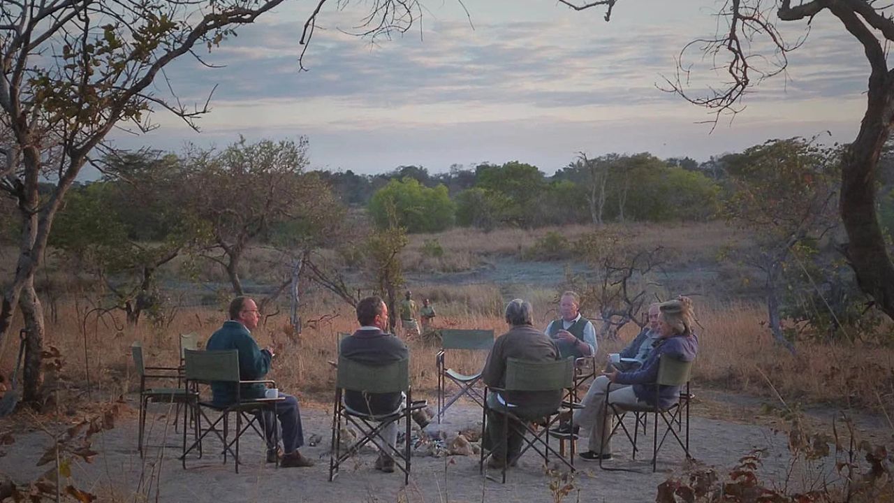Guests seated around a campfire at Musekese Camp with river and bush in background.
