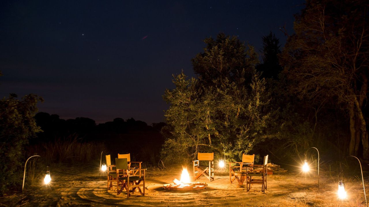 Nighttime Boma campfire glowing in Zambian bush, guests gathered for stories.