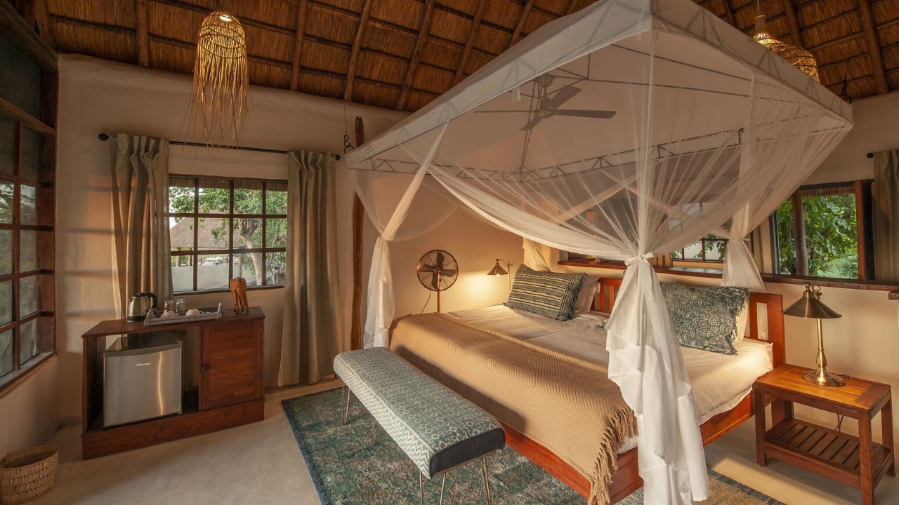 Interior of guest room with double bed, side view, mosquito netting, looking into the bed. Description: The rooms offer cozy interiors with classic safari style, ensuring comfort during stays.