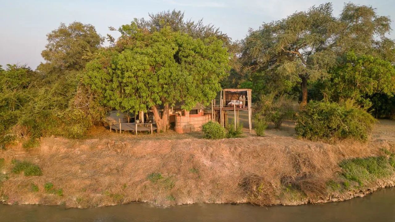 Front view of Three Rivers Camp guest lodge, river in foreground, bush in background, stilted accommodation