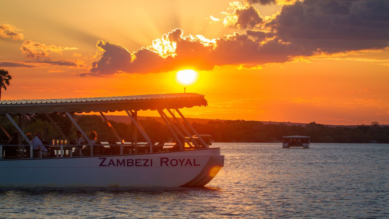 Zambezi Royal boat floating past orange sunset skies on Zambezi River.